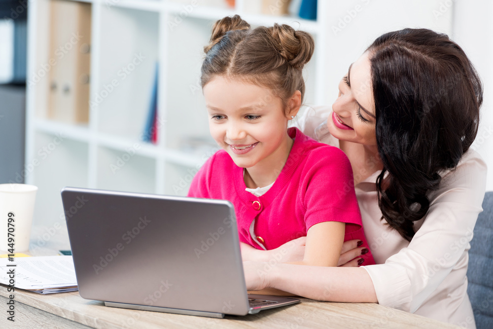 Beautiful happy mother and daughter using laptop at workplace