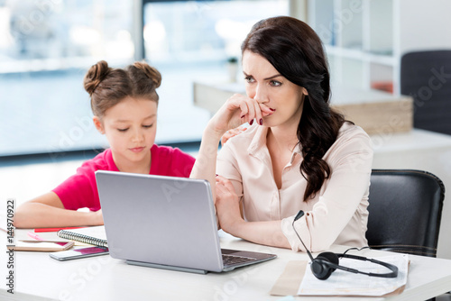 Little girl sitting near upset mother working with laptop and headset at workplace