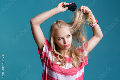 young attractive blond woman brushing her hair with pink comb on blue background