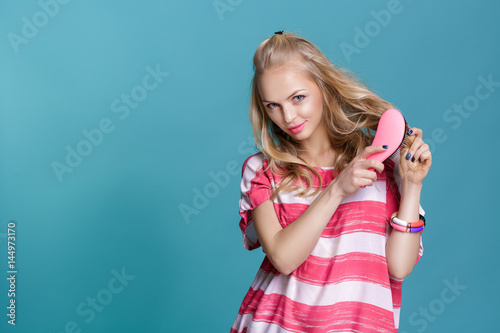 young attractive blond woman brushing her hair with pink comb on blue background