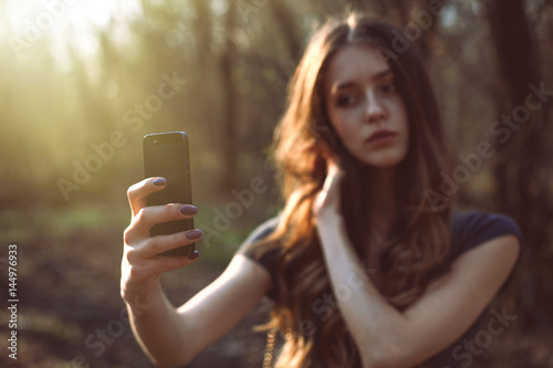 Girl making selfie photo on smartphone in the park. Focus on phone