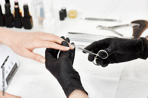 Professional manicurist in black gloves cutting cuticle with manicure scissors.