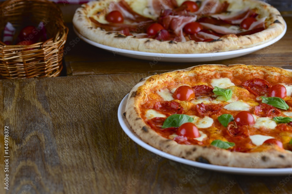 Two hot tasty pizza with pepperoni, prosciutto, mozzarella, cherry tomatoes and basil, served on a dark wooden table for a dinner in a restaurant. Italy food concept. Two hot tasty pizza with pepperoni, prosciutto, mozzarella, cherry tomatoes and basil, served on a dark wooden table for a dinner in a restaurant. Italy food concept.