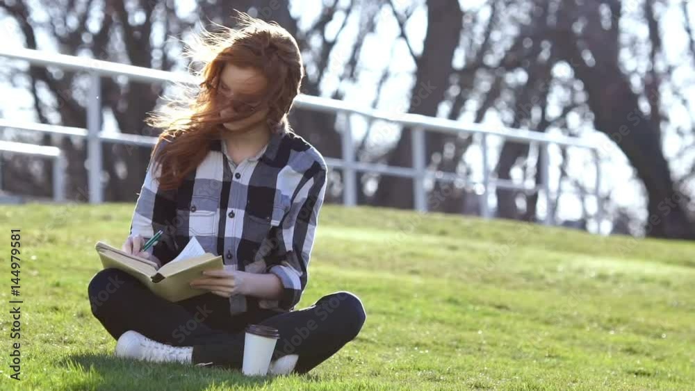 Young pretty woman in sunglasses and shirt sitting in park, writing something and after that reading book Young pretty woman in sunglasses and shirt sitting in park, writing something and after that reading book