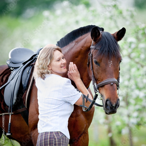 Woman and bay horse in apple garden. Portrait of horse and beautiful lady. Horse rider. Equestrian sport.