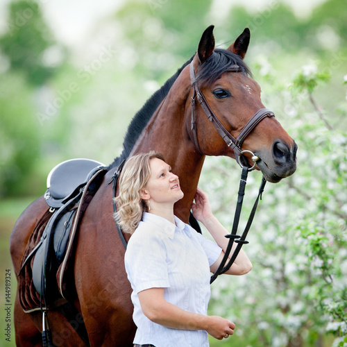 Woman and bay horse in apple garden. Portrait of horse and beautiful lady. Horse rider. Equestrian sport.