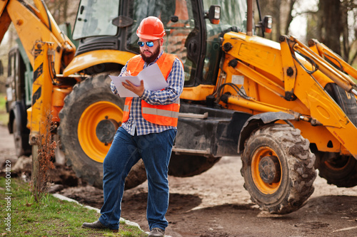 Brutal beard worker man suit construction worker in safety orange helmet, sunglasses against traktor with plan paper at hands.