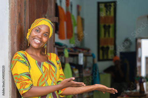 Smiling black African woman in traditional clothes wishing welcome to customers in front of her store