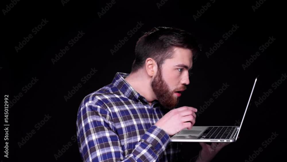 Young surprised bearded man working on laptop while standing isolated over black 