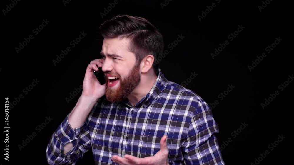 Angry annoyed bearded man shouting while talking on mobile phone isolated over black Angry annoyed bearded man shouting while talking on mobile phone isolated over black