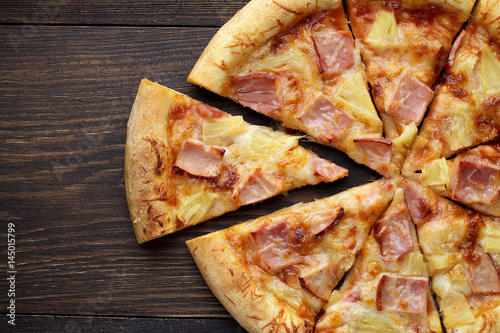 Sliced hawaiian pizza with ham and pineapple on dark wooden table.