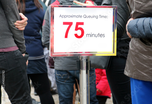 People waiting in a long queue, focus on the information sign. Billboard with information about the time waiting beside line with many people waiting.