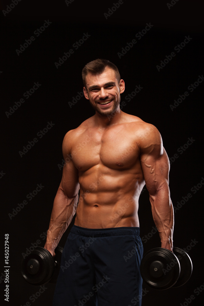 Handsome athletic man in gym is pumping up muscles with dumbbells in a gym. Fitness muscular body isolated on dark background. Handsome athletic man in gym is pumping up muscles with dumbbells in a gym. Fitness muscular body isolated on dark background.
