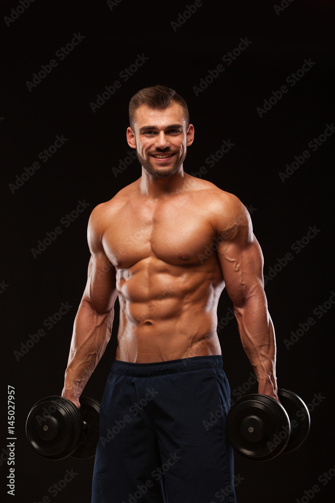 Handsome athletic man in gym is pumping up muscles with dumbbells in a gym. Fitness muscular body isolated on dark background.