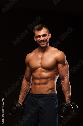 Handsome athletic man in gym is pumping up muscles with dumbbells in a gym. Fitness muscular body isolated on dark background.