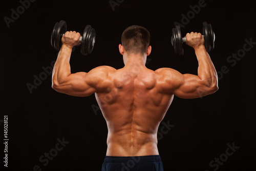 Muscular male model bodybuilder doing exercises with dumbbells, turned back. Isolated on black background with copyspace
