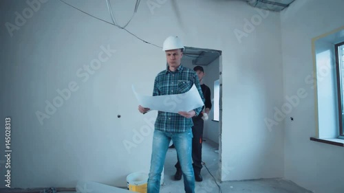 A taskmaster walking on a construction site with the plan then he stop and looking on camera and smile