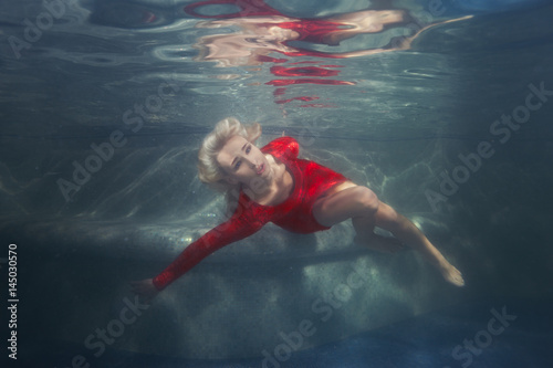 Blond woman under the water in the pool, wearing a red dress.