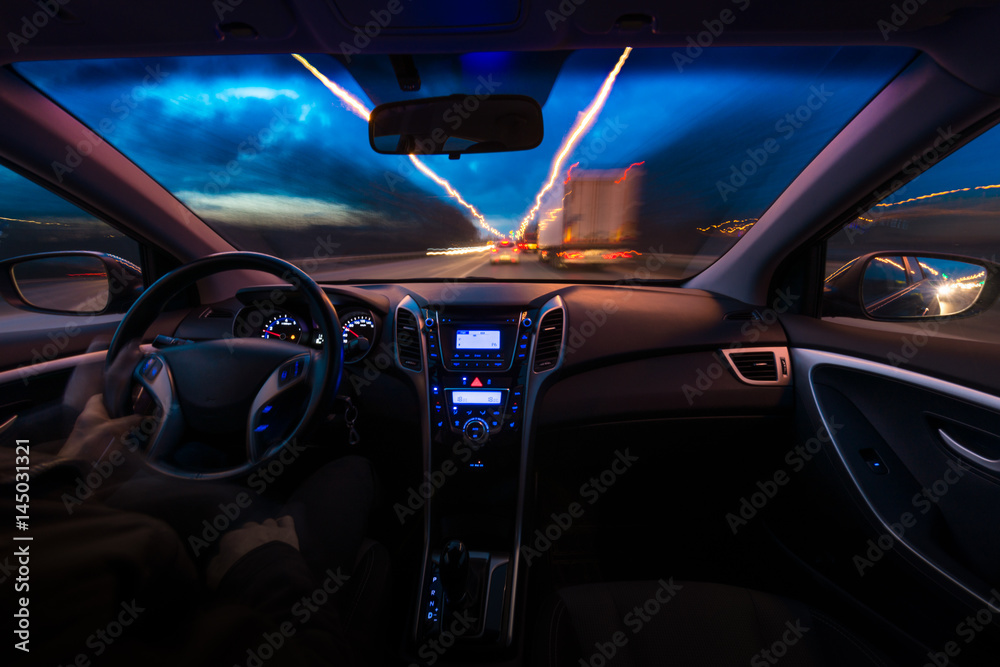 Night view of the road from inside the car. A natural light street and other cars are blurred. The hands of an elderly man