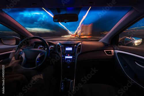 Night view of the road from inside the car. A natural light street and other cars are blurred. The hands of an elderly man
