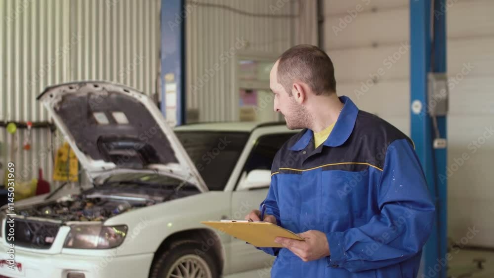 Portrait of a car mechanic writing in a note pad clipboard behind car with opened hood in a repair car service Portrait of a car mechanic writing in a note pad clipboard behind car with opened hood in a repair car service