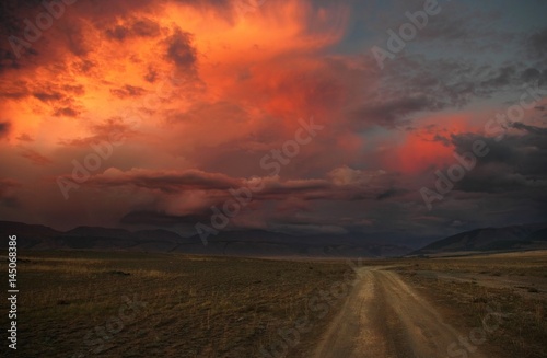 Road path on a desert wild mountain plateau at the background of the hills under a dramatic sunset colorful sky with illuminated red pink purple clouds Kurai Altai Mountains Siberia Russia