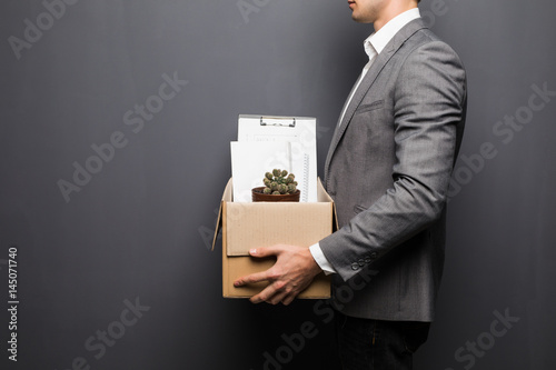 Close up of fired man employee hiding behind box with personal items on grey background photo