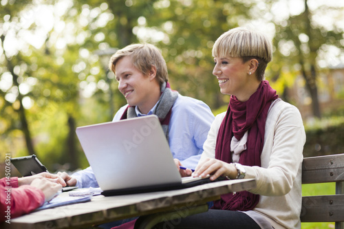 Happy young students sitting with laptop at table in campus photo