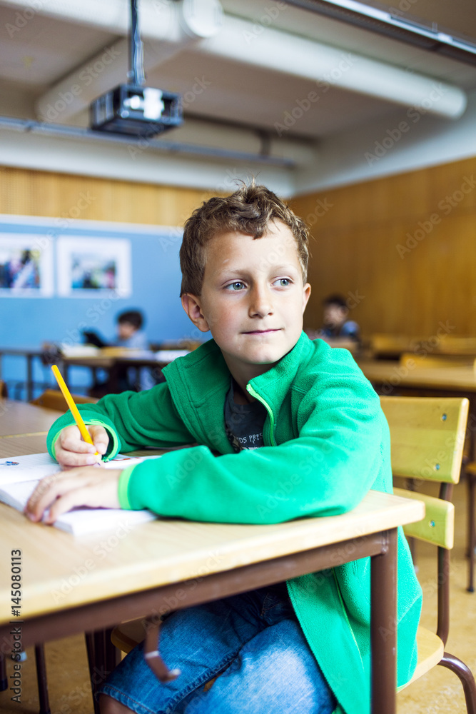 Thoughtful schoolboy looking away while sitting at desk in classroom Thoughtful schoolboy looking away while sitting at desk in classroom