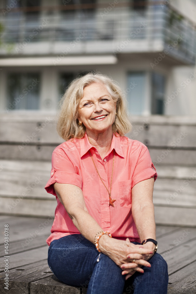 Thoughtful smiling senior woman sitting on wood against building