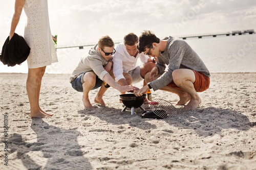 Young male friends lighting barbeque on sandy beach photo