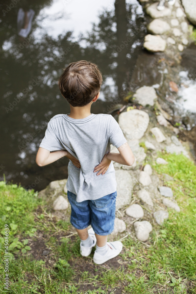 Rear view of boy standing with hands on hip by pond at park Rear view of boy standing with hands on hip by pond at park