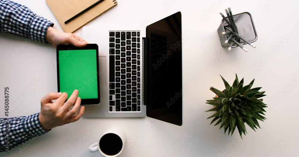 Man working on laptop and black tablet device with green screen on wite