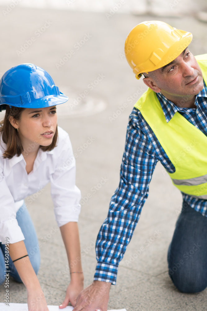 Senior male engineer and young female trainee with safety jackets and hardhats crouching reviewing office blueprints Senior male engineer and young female trainee with safety jackets and hardhats crouching reviewing office blueprints
