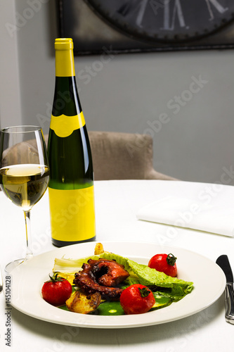 Seafood dish with fried octopus tentacles and vegetables with a bottle and a glass of wine on a white tablecloth background, studio light