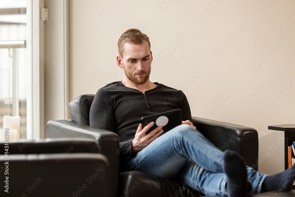 Casual laid back man wearing a black sweater and jeans, sitting down in a lounge chair indoors, holding a digital tablet. Casual laid back man wearing a black sweater and jeans, sitting down in a lounge chair indoors, holding a digital tablet.