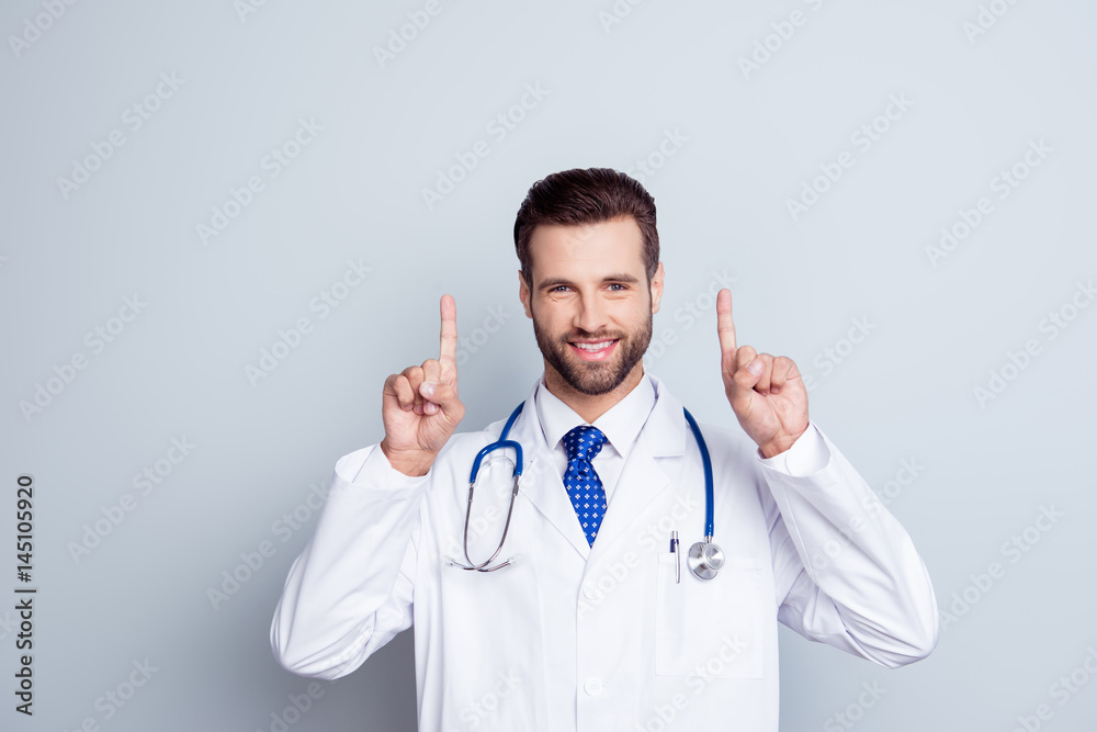 Smiling happy doctor in white coat with stethoscope isolated on gray background pointing up Smiling happy doctor in white coat with stethoscope isolated on gray background pointing up
