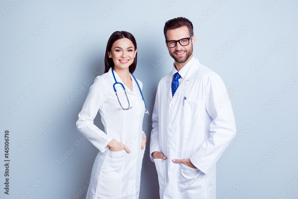 Two best smart professional smiling doctors workers in white coats holding their hands in pockets and together standing against gray background Two best smart professional smiling doctors workers in white coats holding their hands in pockets and together standing against gray background