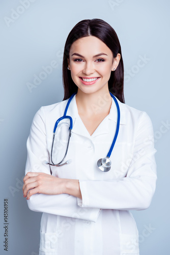 Vertical portrait of charming smiling young female doctor in uniform on gray backgroung standing with crossed hands