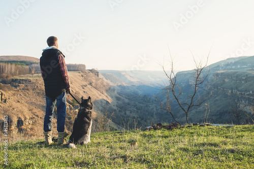 A man with a beard walking his dog in the nature, standing with a backlight at the rising sun, casting a warm glow and long shadows against the background of the gorge and trees.