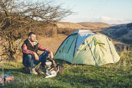 Guy bearded in jeans checkered shirt and a sleeveless jacket with a dog husk on vacation sitting on nature next to the tent in the evening at sunset