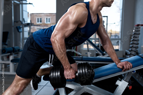 Young muscular man in a T-shirt trains with dumbbells standing with one foot on a bench in the gym close-up