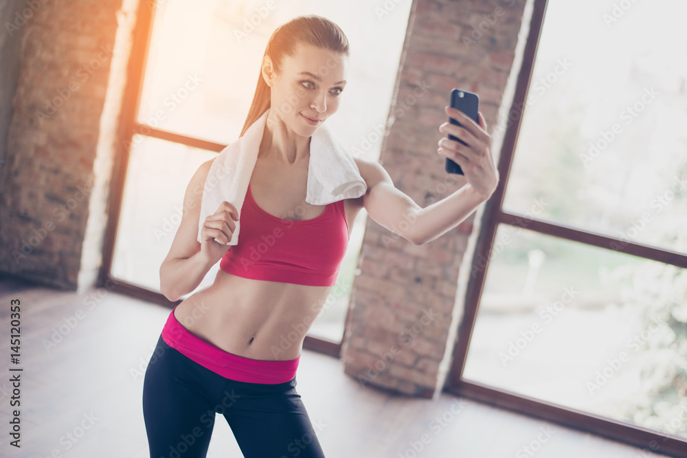 Attractive fit sportgirl is making selfie on her smartphone`s camera after work out. She is smiling, has a towel on shoulders and stylish sportswear