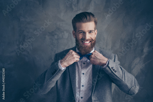 Happy young happy bearded man with mustache in formalewear stands on a gray background and holds his bow-tie with both hands