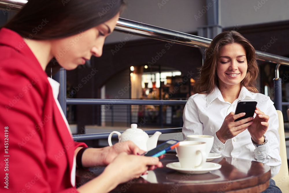Smiling chestnut and serious brunette surfing on Internet in smartphones Smiling chestnut and serious brunette surfing on Internet in smartphones