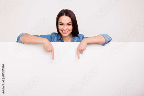 Cheerful cute girl is standing behind the white blank banner and pointing down at a copyspace photo