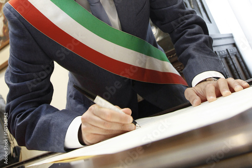 foto b/n della mano di un sindaco con fascia tricolore che firma con una penna un documento photo