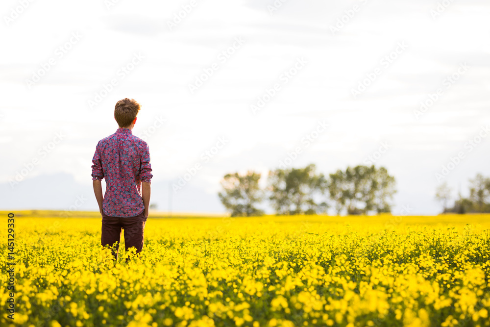 Young Man Standing In Canola Field Looking At Mountains In Summer