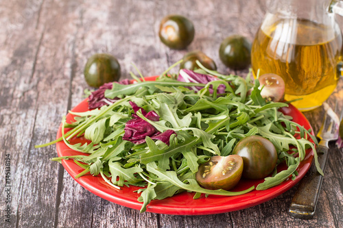  Green salad. Fresh green salad with tomatoes on a red plate, next to a glass jug with olive oil.