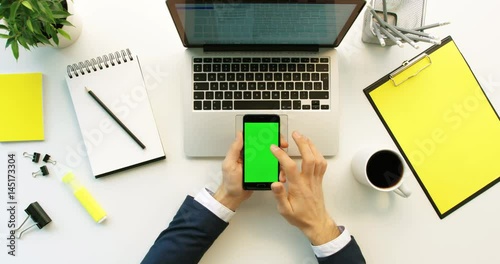 Business man using smart phone with green screen, scrolling pages, tapping on touch screen on office desk background. Hands top view. Green screen. Chroma key photo